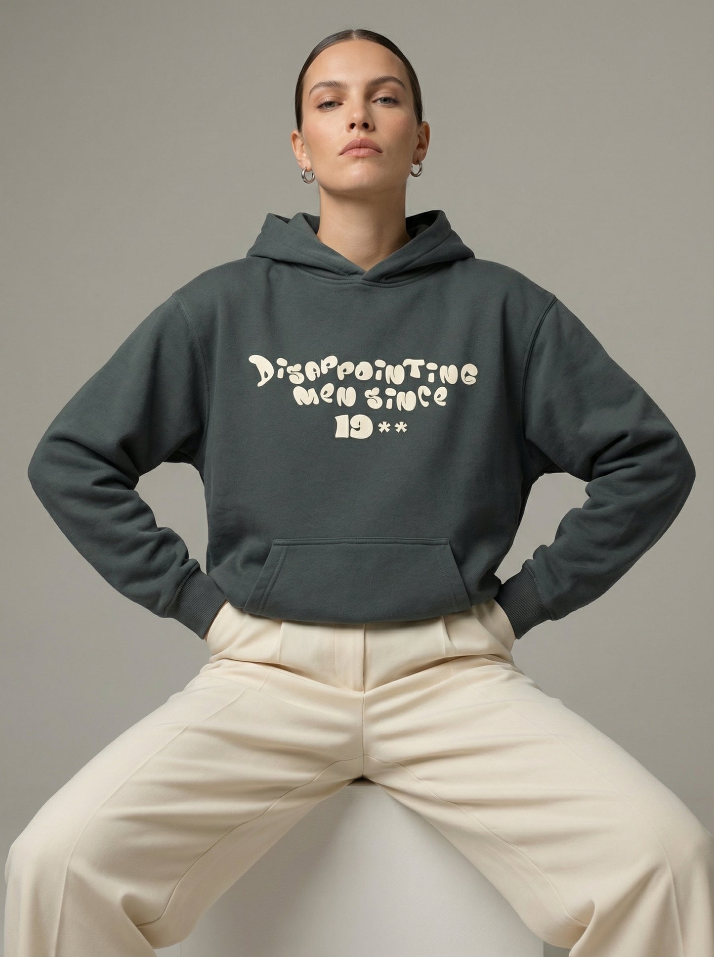 Person wearing a dark gray hoodie with text"Disappointing Men Since 19**", sitting on a white stool against a neutral background