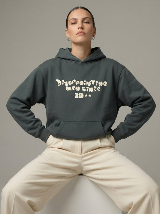 Person wearing a dark gray hoodie with text"Disappointing Men Since 19**", sitting on a white stool against a neutral background