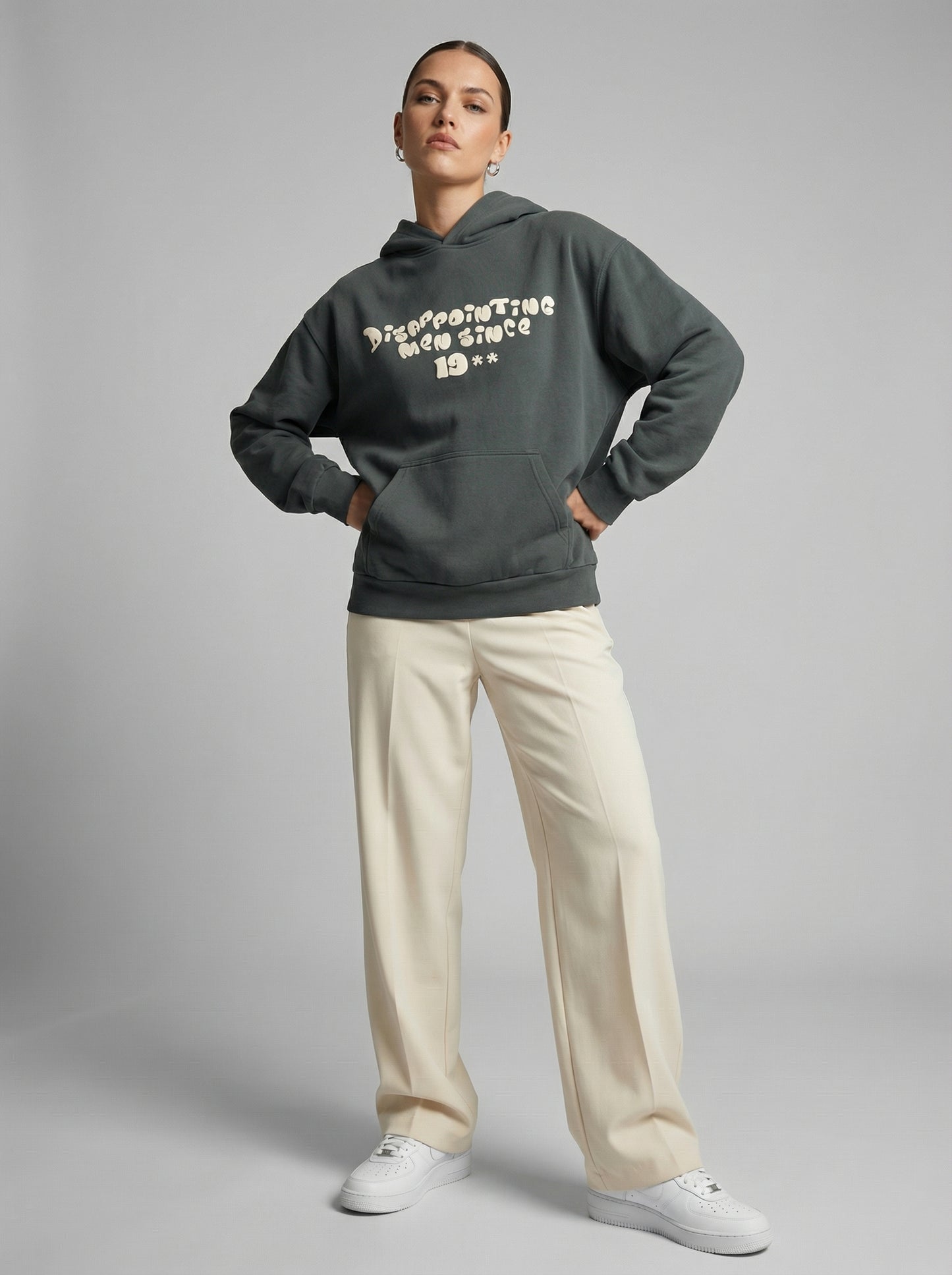 Person wearing a dark gray hoodie with text "Disappointing Men Since 19**", beige pants, and white shoes on a gray background