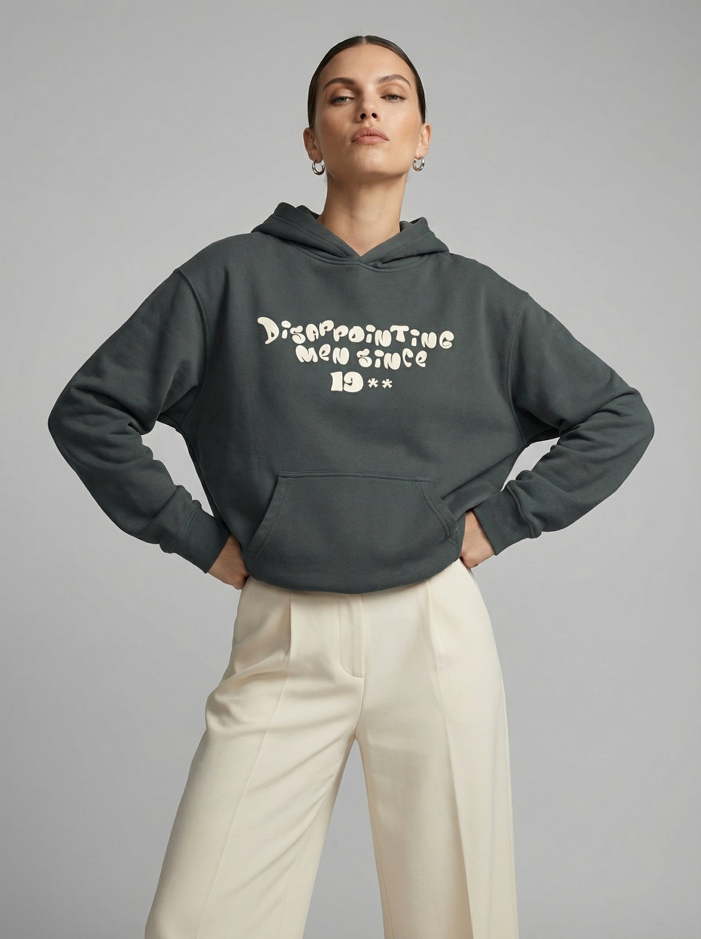 Person wearing a dark gray hoodie with text "Disappointing Men Since 19**" and light-colored pants on a gray background