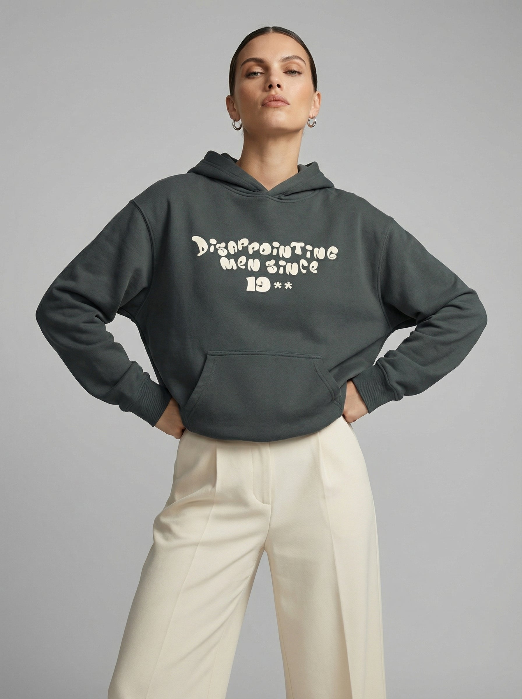 Person wearing a dark gray hoodie with text "Disappointing Men Since 19**" and light-colored pants on a gray background